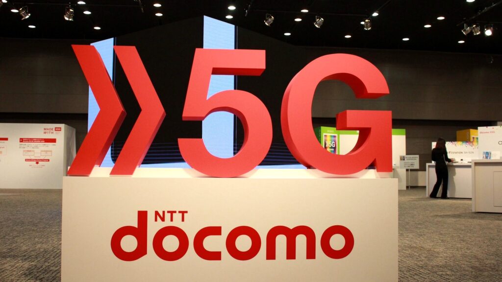赤い立体の5Gの文字とNTT docomoのロゴの奥に人物や案内板が見える展示会場の写真。「料金変更」に4.7秒で聞き返すドコモのAIは3往復目で同じ質問に戻る