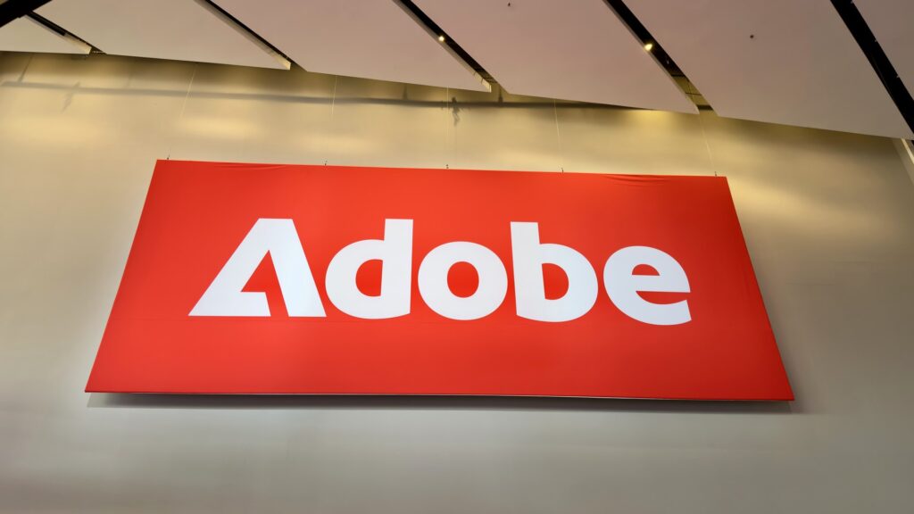 白い天井パネルの下につり下げられた、赤地に白いAdobeのロゴが入った大きな看板の写真。Adobe、GeminiでPhotoshopを0円で使える 日本語は未対応だ