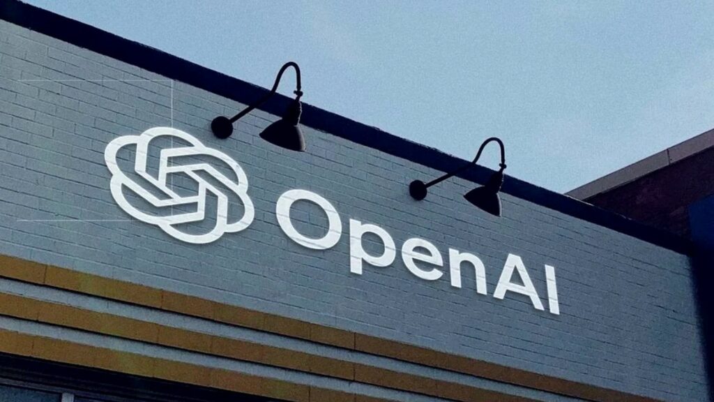 灰色のレンガ壁にOpenAIのロゴマークと社名が白く描かれ、上部に壁付けのアームライトが並ぶ建物外観の写真。ChatGPTで仕事と個人の切り替えが3つから全プラグインに広がった