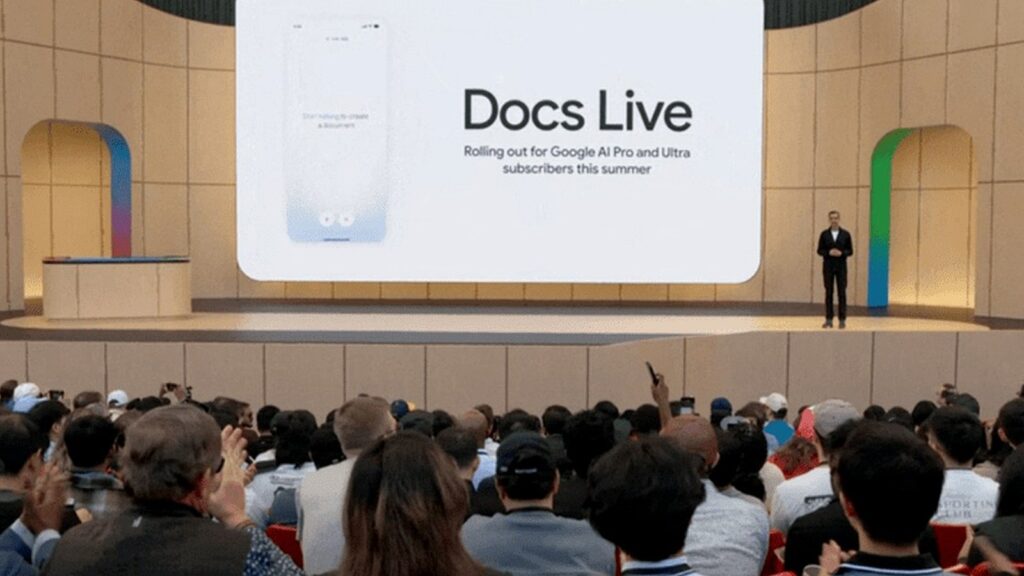 Google I/Oカンファレンスの舞台上に「Docs Live」というテキストが表示される大型スクリーンがあり、その下で男性が立っている。観客が大勢座っており、スマートフォンで撮影している姿が見える。舞台両脇にはカラフルなアーチ装飾がある。