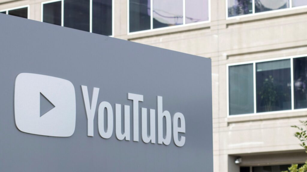 YouTubeのロゴ（再生ボタン）と社名が白く浮き出たグレーの屋外看板と、背後に見えるガラス窓のあるビルの写真。YouTube売上2年で13倍でもGemini編集AIは日本未定だ