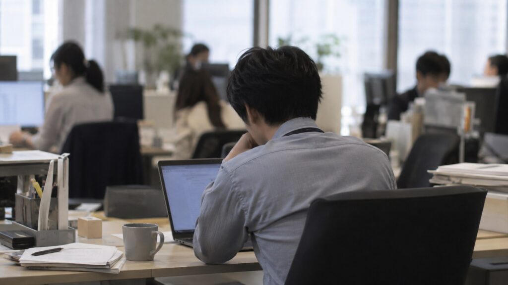 日本のオフィスの席でノートパソコンの画面を見ながら手を止めて考えている会社員の後ろ姿。机にはマグカップと書類、奥に同僚が数人いる