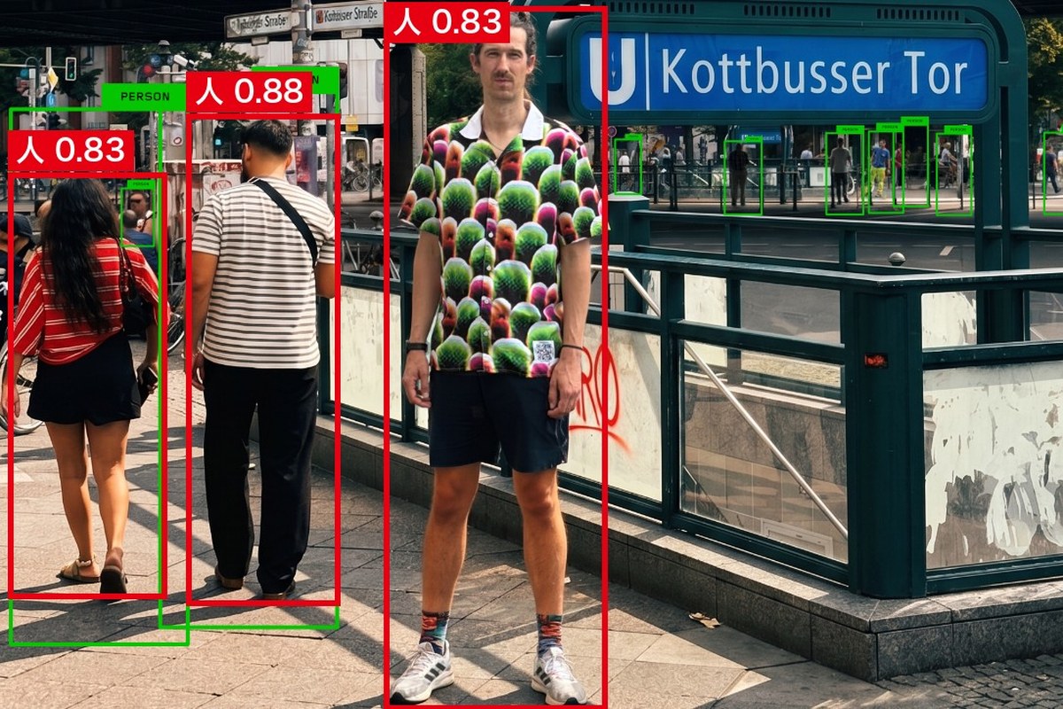 駅の入口の前で迷彩柄のシャツを着た人が立つ写真。作者が付けた緑の枠は左の2人にだけあり、AInformationがYOLO11nで判定した赤い枠はシャツの人にも付いて「人 0.83」の表示が出ている