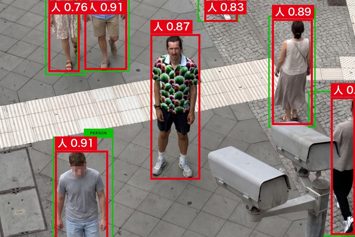 監視カメラ越しに通りを上から撮った写真。作者の緑の枠はシャツの人に付いていないが、AInformationがYOLO11nで判定した赤い枠はシャツの人を囲み「人 0.87」の表示が出ている
