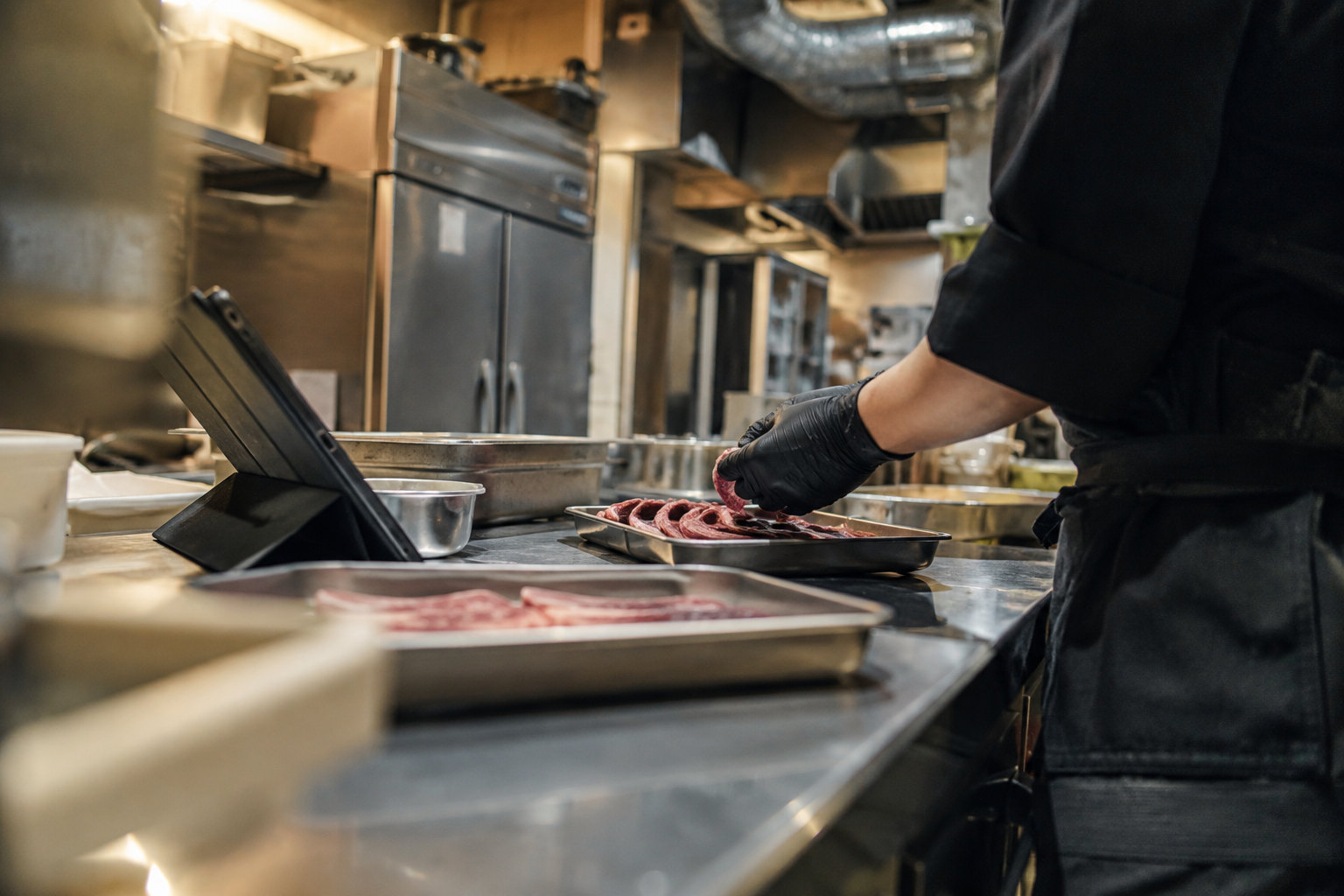 焼肉店の厨房。ステンレスの作業台に肉のトレーとタブレット。奥に冷蔵庫と換気のダクト。調理スタッフの手元だけ。厨房の照明