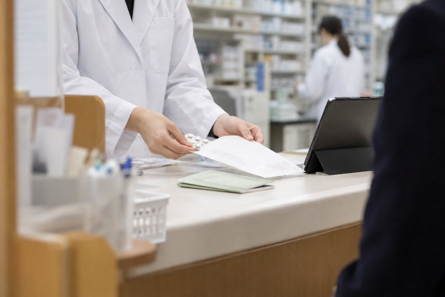 調剤薬局の投薬カウンター。薬袋とお薬手帳、その横にタブレット。奥に調剤室の棚が見える。薬剤師の手元だけ。明るい室内