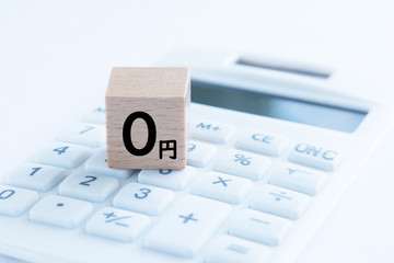 明るい水色がかった背景で、白い電卓のボタンの上に「0円」と書かれた木のブロックが置かれた写真
