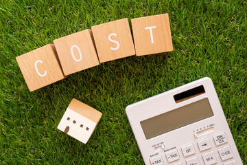 緑の芝生の上に、「COST」の文字が入った木のブロックと小さな家の形のブロックと電卓が置かれた写真