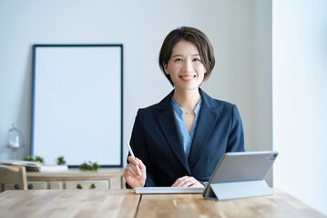 明るい部屋の木の机で、紺のジャケット姿の女性がタブレットの前に座って微笑んでいる写真