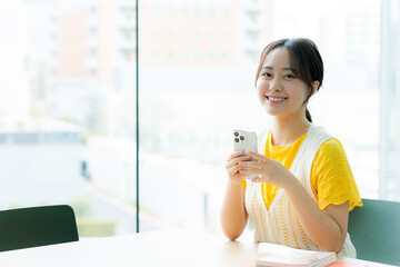 大きな窓の前のテーブルで、黄色い服に白いベストの女性がスマホを両手で持って微笑んでいる写真
