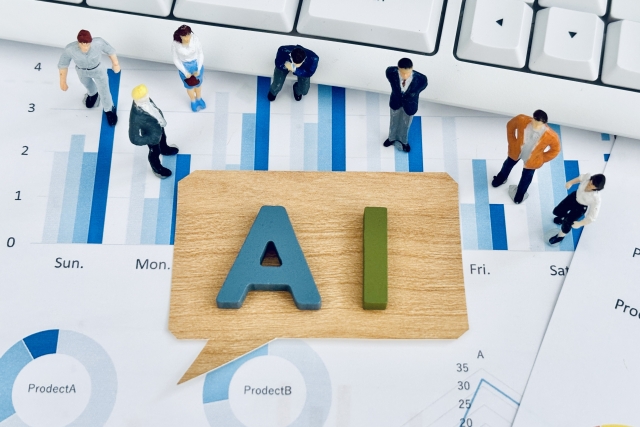 AIの文字を載せた木の吹き出しのまわりにビジネスパーソンの人形が立つ、グラフの資料の上を写した写真