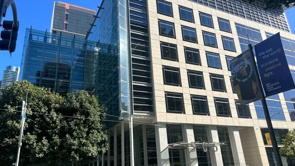 青空の下、ガラス張りと石張りのオフィスビルが並ぶ交差点を見上げ、手前に街路樹と信号機が写る写真。Anthropic、技術職の若手採用が27%から9%に下がった 会議は増えた