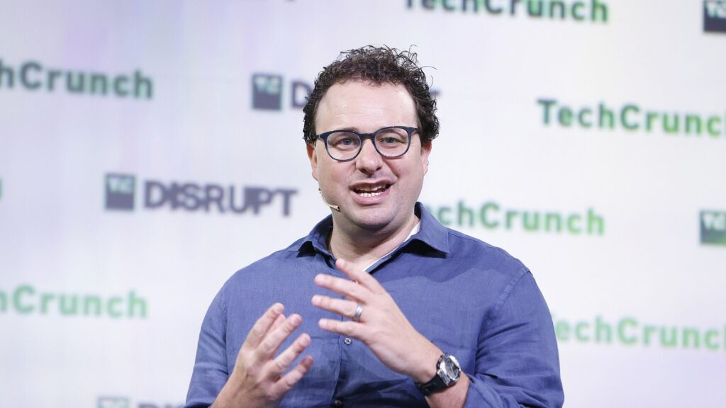 TechCrunch Disruptのロゴが並ぶ壇上でメガネをかけた男性が身振りを交えて話している写真。Anthropic、Claudeの文章に透かし 追加0円で一般は確かめられない