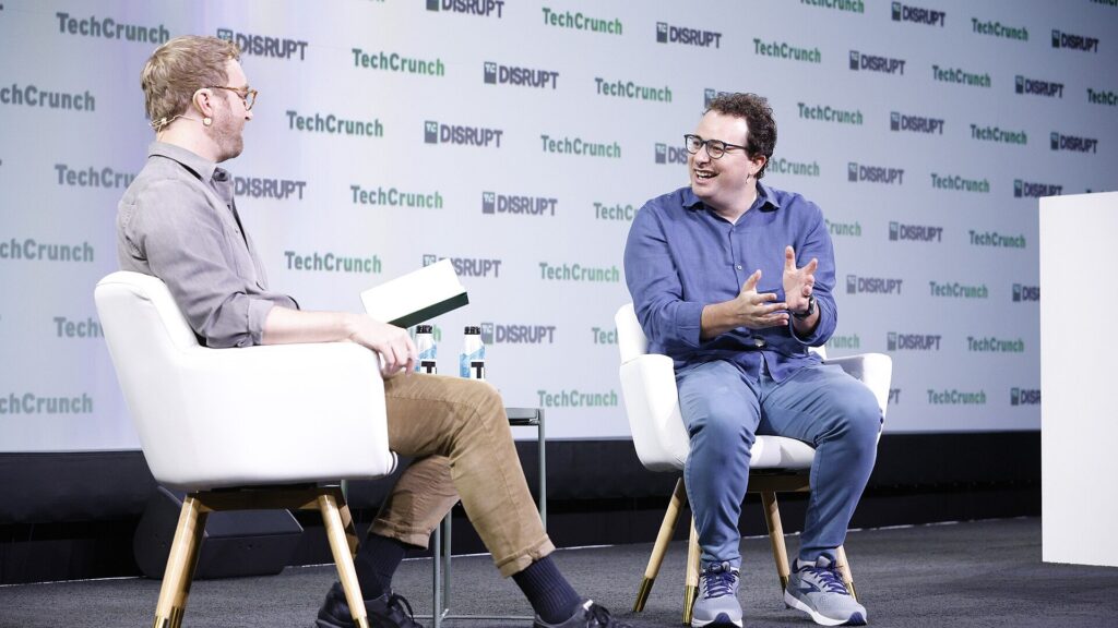 TechCrunchのロゴが並ぶ背景の前で白い椅子に座った男性2人が向き合って談笑しているステージ写真。Geiger、AIエージェントを0.26秒で一覧 スキルは50本で切れる