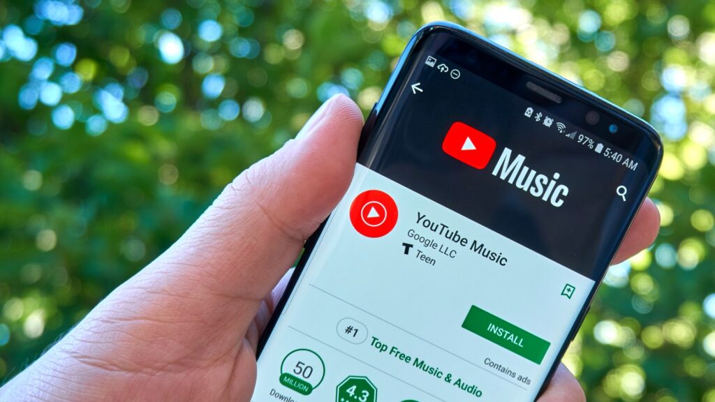 手元がスマートフォンを持ち、Google PlayストアでYouTube Musicアプリのインストール画面を表示している写真。YouTube、AI曲に表示を開始 3区分を申告しなくても検出で付く