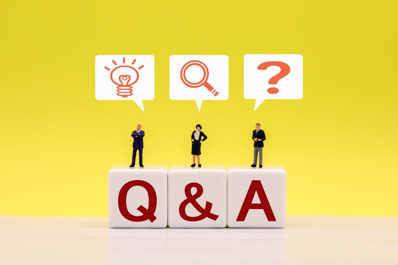 黄色い背景の前で「Q&A」の文字のキューブの上に3体のミニチュア人形が立ち、上に電球と虫眼鏡とクエスチョンマークの吹き出しが並んだ写真