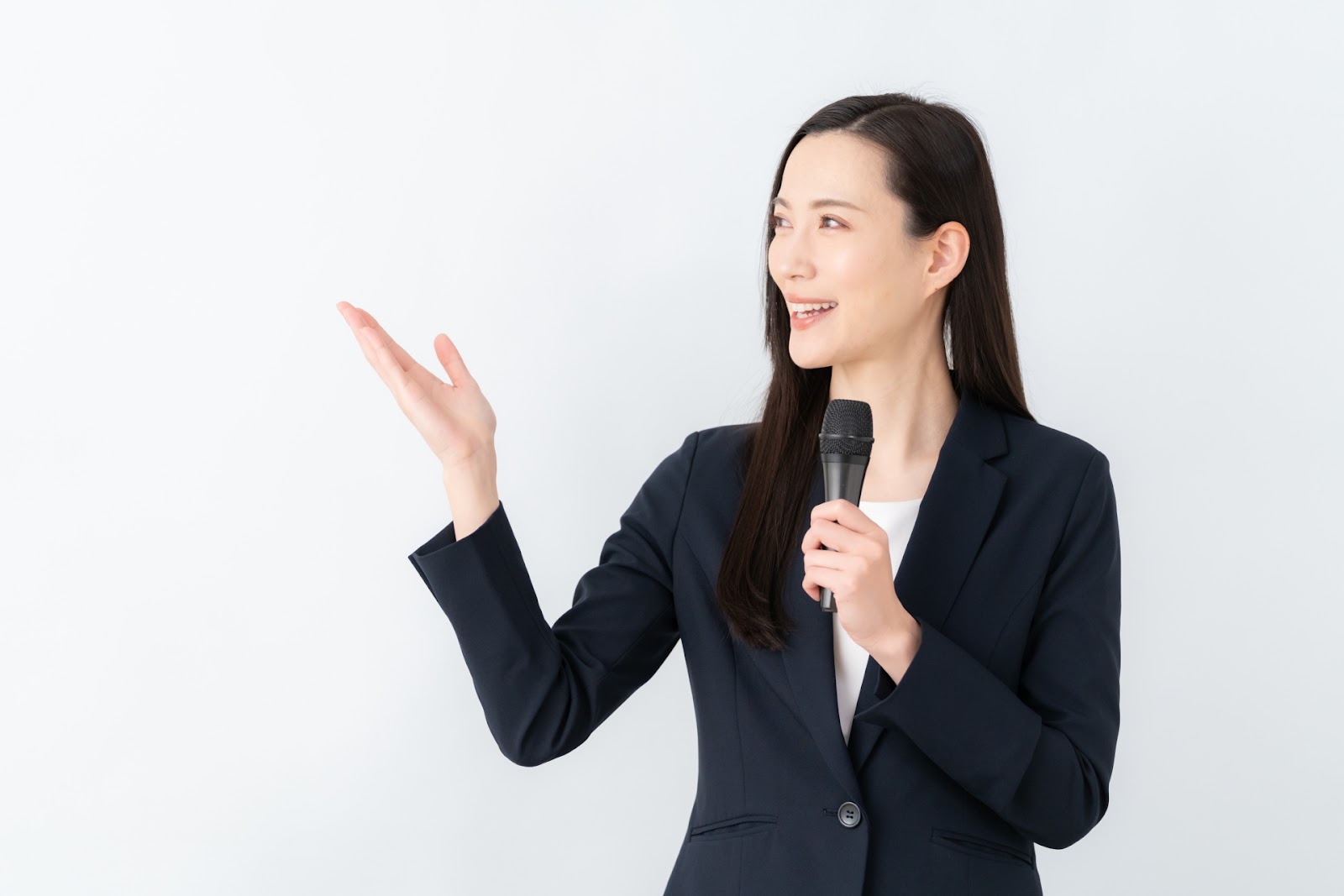黒いスーツの女性が白い背景の前でマイクを持ち、片手を横に広げて笑顔で話しているイメージ写真