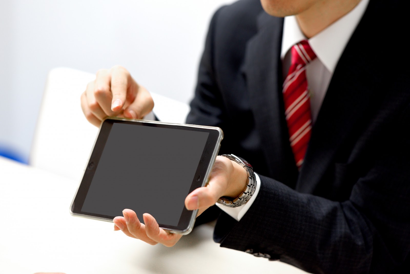 赤いストライプのネクタイを締めたスーツの男性が、画面の黒いタブレットを手前に向けて見せている写真