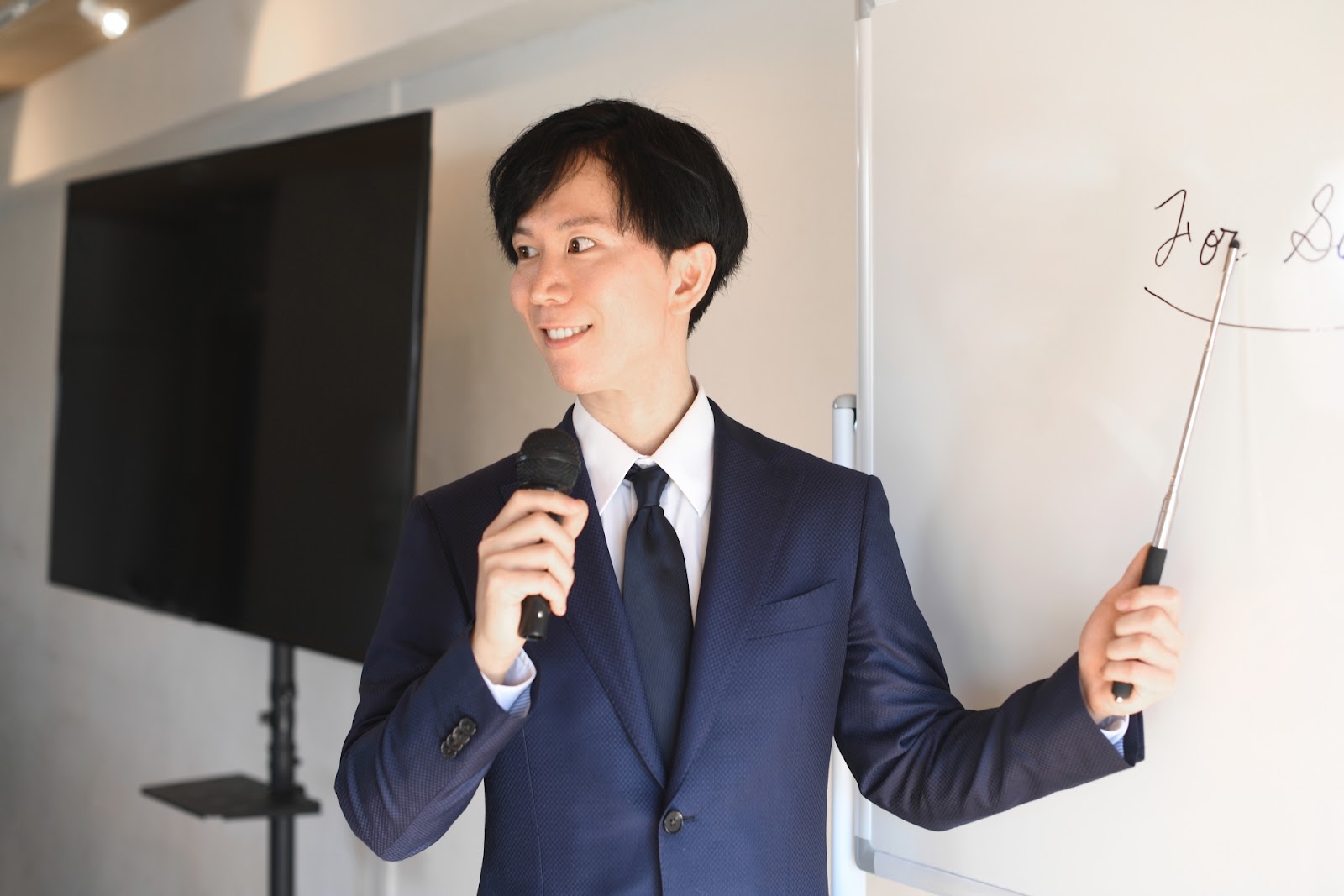 紺のスーツの男性がマイクと指し棒を持ち、ホワイトボードとモニターの前で講義をしている写真