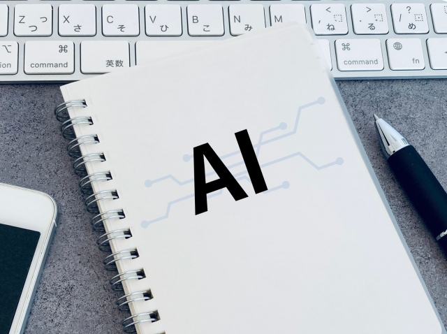 灰色の机の上で、「AI」と大きく書かれた白いノートがキーボードとスマホとペンの間に置かれた写真