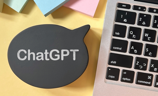 「ChatGPT」の文字が入った黒い吹き出しの置物が、付箋とキーボードのある黄色い机の上に置かれた写真
