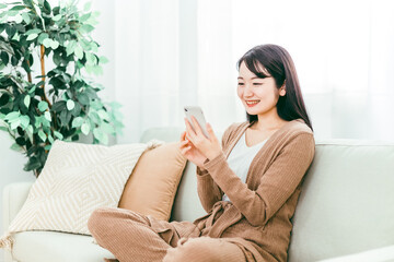 観葉植物と白いカーテンのある明るい部屋で、ソファに座った女性が笑顔でスマホの画面を見ている写真