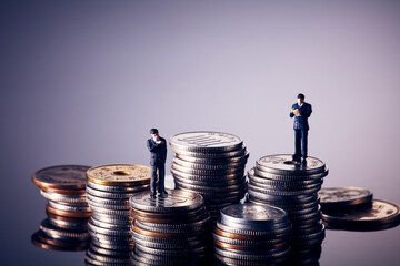 暗い紫色の背景で、積み重ねた硬貨の山の上にスーツ姿のミニチュア人形が2体立っている写真