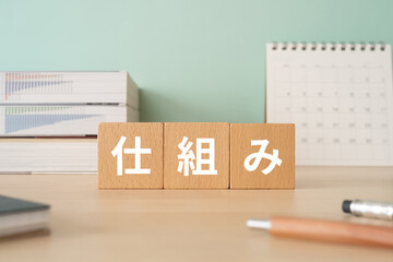 「仕組み」の文字が入った3つの木のブロックが、本やカレンダーやペンのある机の上に並んだ写真
