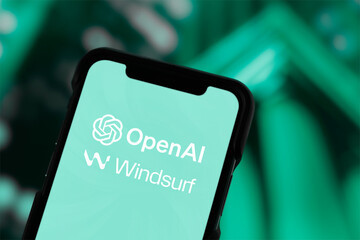 薄い緑色の画面に「OpenAI」と「Windsurf」のロゴを表示したスマホを、濃い緑の背景で写した写真