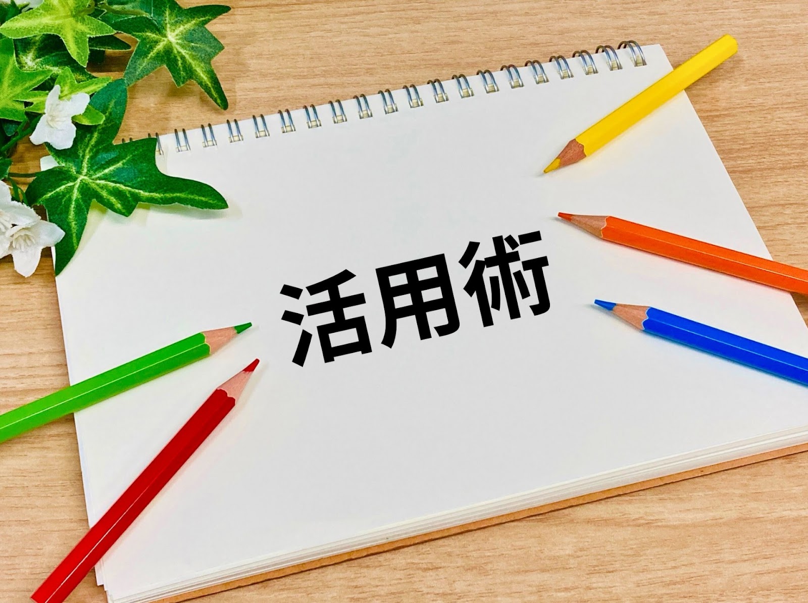 木の机の上の白いノートに大きく「活用術」と書かれ、周りにカラフルな色鉛筆と観葉植物の葉が置かれた写真