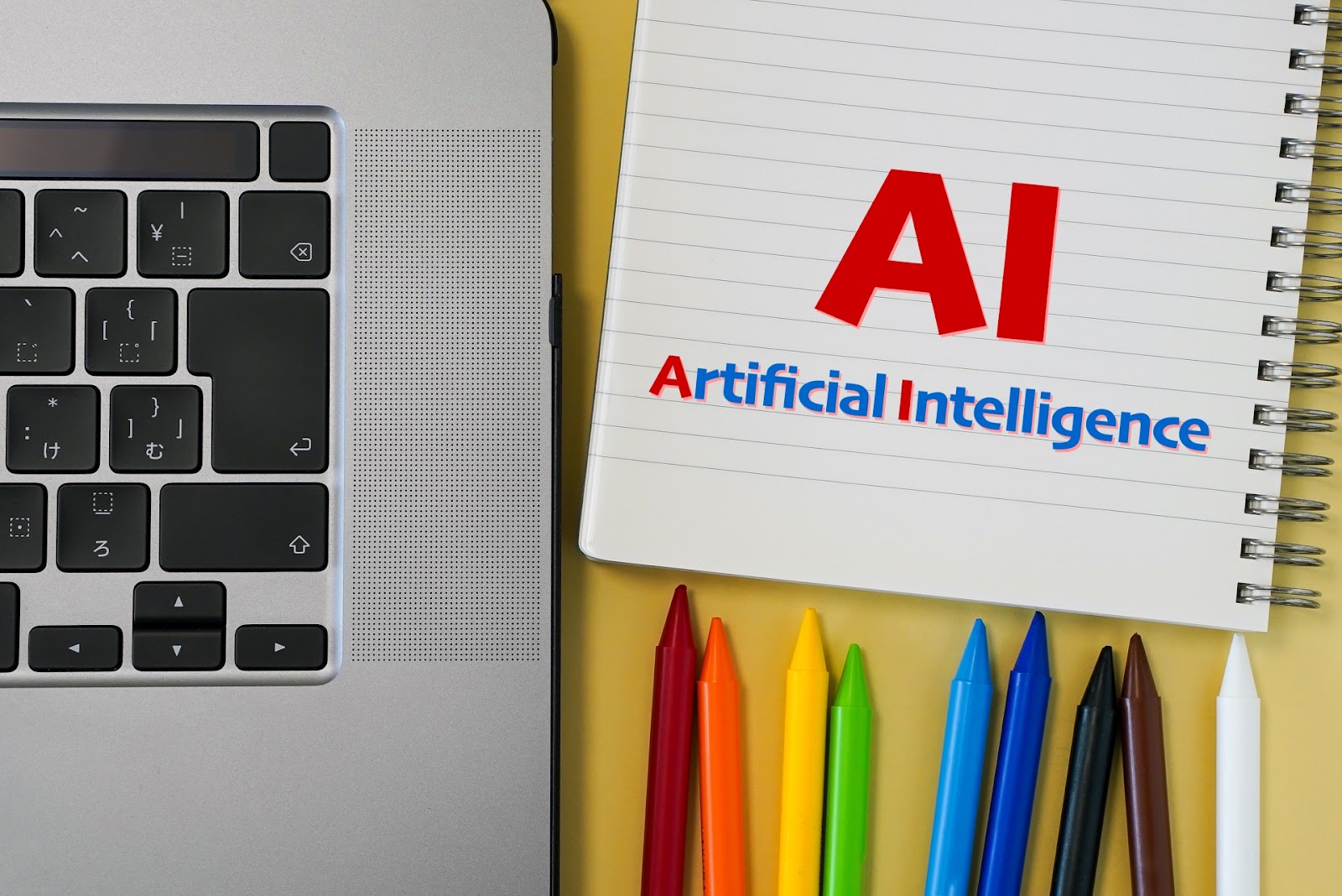 ノートパソコンのキーボードの横に、「AI」と「Artificial Intelligence」の文字が書かれたノートと色鉛筆が並んだ写真