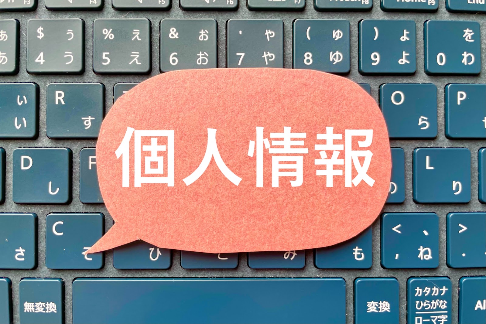 「個人情報」と書いた赤い吹き出しの紙を青いキーボードの上に置いて真上から撮った写真