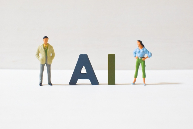 白い台の上で、男性と女性のミニチュア人形が青い「A」と緑の「I」の木の文字を挟んで立つ写真