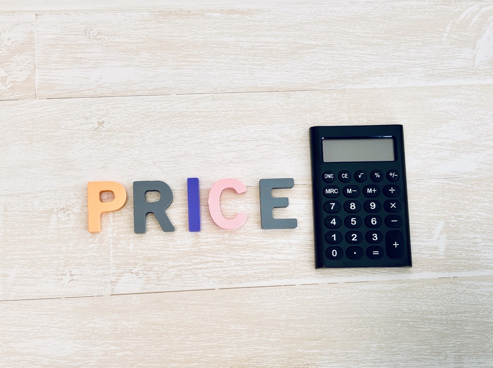 白い木目の板の上に、オレンジやピンクなどカラフルな立体文字の「PRICE」と黒い電卓を横に並べて置いた写真