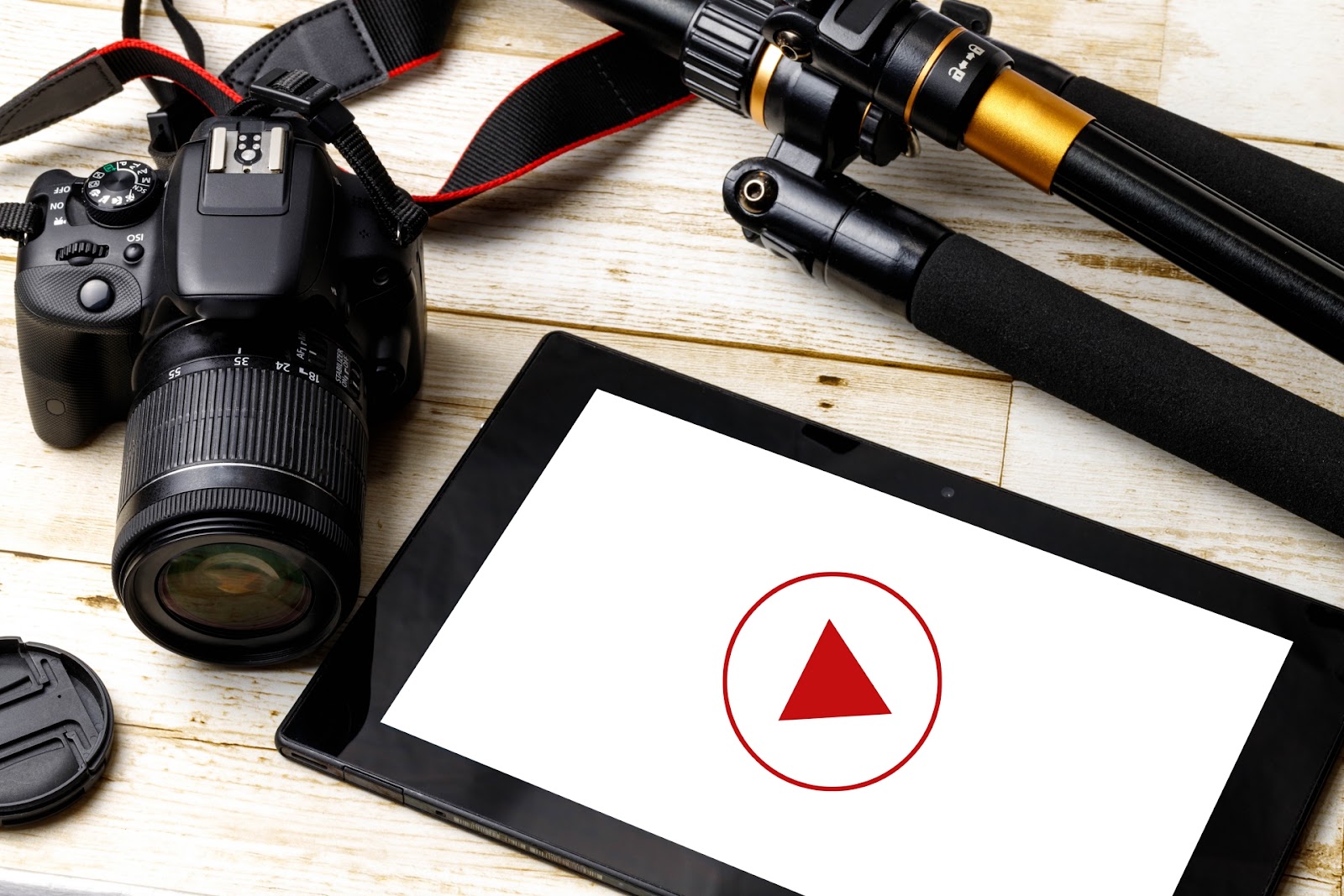 黒い一眼レフカメラと三脚と赤い再生マークが出たタブレットが白い木の机の上に並んだ写真