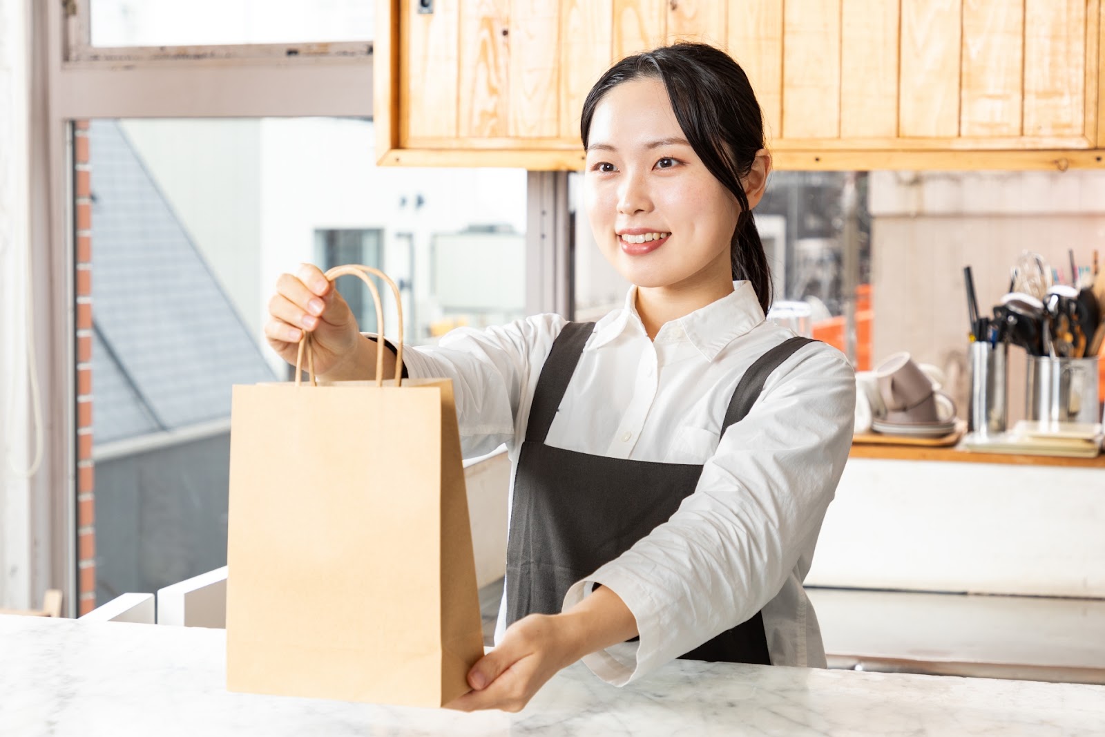 白いシャツにエプロン姿の女性が明るい店内のカウンター越しに茶色の紙袋を笑顔で差し出している写真